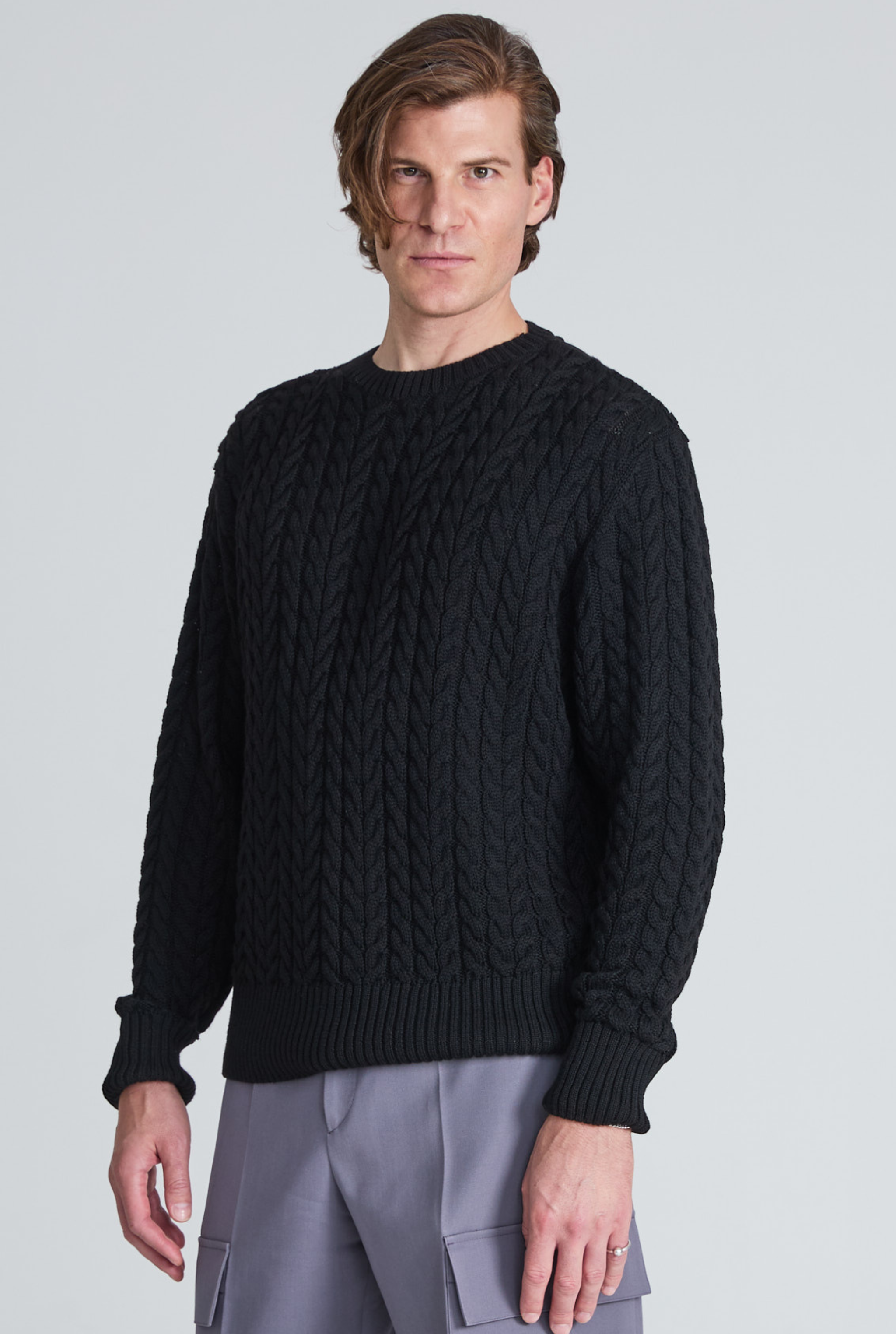 Person klädd i en beige, kabelstickad tröja med handen delvis instoppad i fickan på svarta byxor.